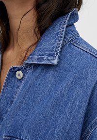 Primer plano de una persona que lleva una chaqueta de mezclilla azul con costuras visibles y un botón de metal cerca del cuello.