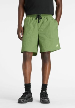 Mann trägt grüne Sportshorts mit schwarzer Kordel, schwarze Socken und schwarze Laufschuhe, steht mit Händen in den Taschen.