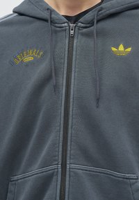 Close-up van een grijze zip-up hoodie met gele Adidas Originals-logo en trefoil-embleem op de borst, voorzien van trekkoorden en zakken aan de voorkant.