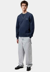 Sudadera azul marino con cuello redondo y detalle de logo, combinada con jeans de pierna ancha gris claro y zapatos negros, de pie contra un fondo blanco.