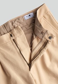 Pantalon beige avec une fermeture éclair, un bouton et des détails de couture discrets. Le tissu semble lisse avec un design structuré.