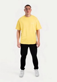 Gul bomulls-T-shirt med rund halsringning och korta ärmar, kombinerad med svarta byxor och vita sneakers. Enkel design i enfärg.