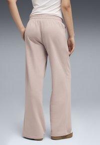 Personne portant un pantalon large rose clair, un haut blanc et des baskets Puma marron, debout avec une main posée sur la hanche.