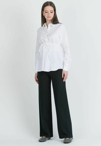 Blusa bianca con bottoni e vita legata, abbinata a pantaloni neri a gamba larga. La parte inferiore ha una leggera lucentezza; le scarpe presentano un design con borchie.