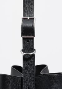 Czarny skórzany pasek torby o teksturowanym wykończeniu. Zawiera srebrną metalową klamrę i pierścień, z widocznymi szwami i regulowanymi otworami.