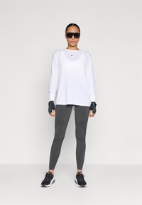 Λευκή μπλούζα με μακριά μανίκια και μαύρο λογότυπο, γκρίζα κολάν με υφασμάτινες πλάκες, μαύρα γάντια και μαύρα αθλητικά παπούτσια με λευκές λεπτομέρειες.