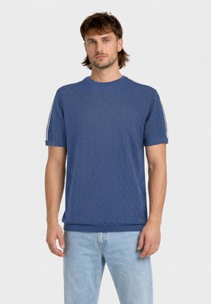 Uomo che indossa una maglia a maniche corte testurizzata blu con strisce bianche sulle maniche e jeans celesti, in piedi contro uno sfondo semplice.