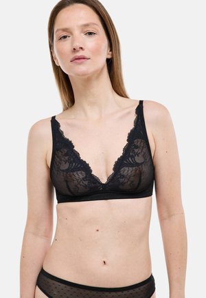 Femme portant un soutien-gorge en dentelle noire et une culotte en maille assortie, debout devant un fond gris clair uni.