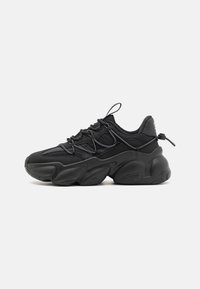 Zapatillas negras con suela gruesa, que presentan una parte superior de malla, cordones detallados y una lengüeta en el talón para ponerlas y quitarlas fácilmente.