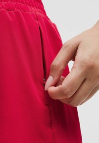 Raudoni sportiniai šortai su šoniniu užtrauktuku. Audinys lygus, matomos siuvimo detalės. Rankos laikosi užtrauktuko traukimo.