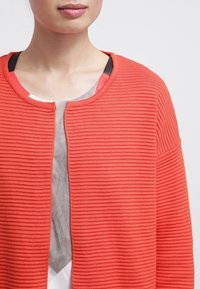 Korallfärgad ribbad cardigan med öppen framsida och rund halsringning. Texturerat tyg och sänkta axlar. Lager på en ljusfärgad topp.