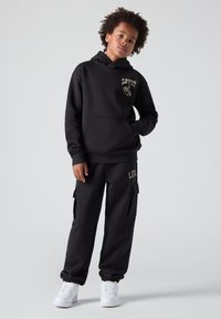 Černá mikina s kapucí a odpovídající cargo kalhoty, obě vyrobené z měkké tkaniny. Obsahuje potisk loga a boční kapsy. Kombinováno s bílými teniskami.