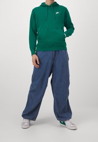 Grön bomulls-hoodie med framficka och dragsko, kombinerad med lösa blå denimbyxor. Har en vit Nike-logga på bröstet.