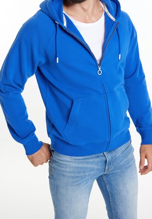 Blue zip-up hoodie made of cotton blend, featuring a front pocket, drawstring hood, and ribbed cuffs. Worn over a white t-shirt and jeans.