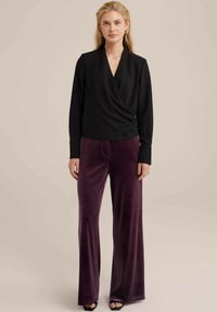 Blusa nera avvolgente con maniche lunghe, abbinata a pantaloni in velluto a vita alta e a gamba larga di un profondo viola. Dettaglio semplice con bottone sulla blusa.
