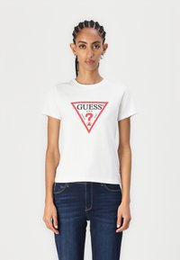 T-shirt en coton blanc avec un logo triangulaire rouge portant "GUESS" et un point d'interrogation, associé à un jean bleu foncé. Manches courtes, col ras du cou.