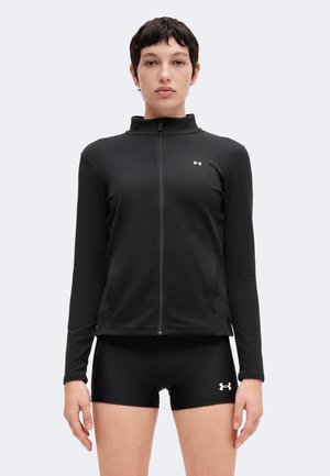Jeune personne aux cheveux courts et foncés portant une veste de sport noire zippée et un short de compression noir avec des logos blancs sur la poitrine et la jambe.