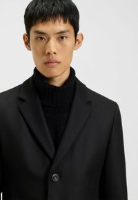 Schwarzer Wollmischmantel mit Reverskragen und einem Knopfverschluss, getragen über einem schwarzen gerippten Rollkragenpullover. Einfarbiger Hintergrund.