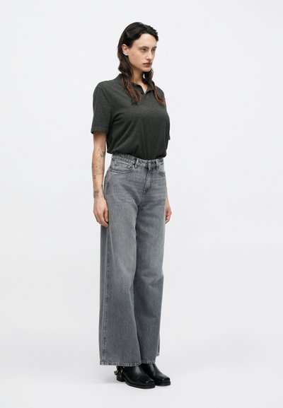 Polo gris, jean large taille haute gris, bottes noires et tatouages sur un bras ; fond blanc uni mettant en valeur les détails des vêtements.