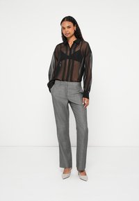 Γυναίκα που φοράει μαύρο διάφανο μπλουζάκι πάνω από μαύρο σουτιέν, γκρι ραμμένο παντελόνι και μπεζ ψηλοτάκουνα, στέκεται μπροστά σε λευκό φόντο.