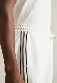 Pantaloni atletici din plasă albă cu o suprafață texturată, având o talie cu șnur și două benzi laterale în negru și bej.