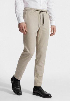 Man draagt beige broek met trekkoord, zwarte leren loafers en een wit overhemd met lange mouwen, getoond van taille tot voeten lopend.