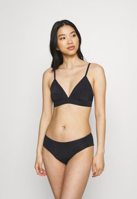 Mulher com cabelo longo e escuro vestindo um conjunto de lingerie preto de duas peças, olhando de lado contra um fundo claro e simples.