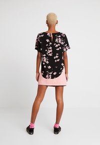 Person med kort blont hår som har på sig en svart blommig skjorta, rosa kjol, rosa strumpor och svarta skor, vänd bort mot en plain vit bakgrund.