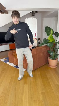 Fekete, hosszú ujjú póló "Ngagani" felirattal, világoskék nadrág és ezüst tornacipő. Zárt tér, barna kordbársony kanapéval és nagy növénnyel.