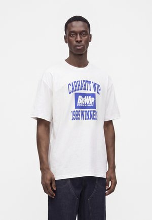 Mand iført en hvid Carhartt WIP t-shirt med blå "Big WIP Conference 1989 Winners" tekst, stående foran en ensfarvet hvid baggrund.