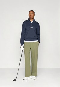 Granatowy pulower na suwak do ćwiartki z wyhaftowanym logo, zestawiony z oliwkowozielonymi szerokimi spodniami i białymi butami, trzymający kij do golfa.