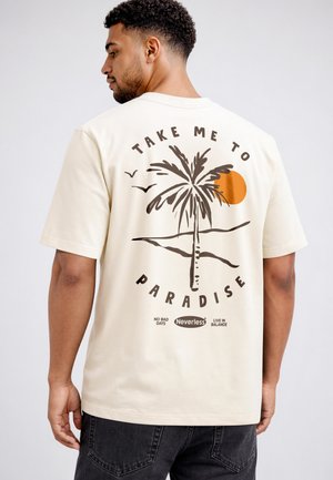 Man wearing cream t-shirt with palm tree, orange sun, birds graphic, and text "Take me to paradise," "No bad days," "Nevertheless," "Live in balance."