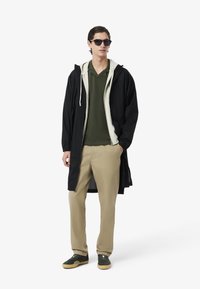 Lång svart jacka över en krämfärgad hoodie, grön polo, beige byxor och gröna sneakers med gummisula; solglasögon fulländar outfiten.