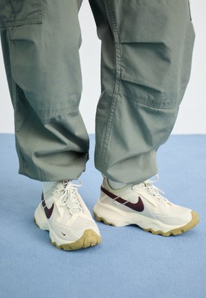 Białe i beżowe sneakersy Nike z ciemnym logo Swoosh, noszone z luźnymi oliwkowymi spodniami typu cargo na jasnoniebieskiej podłodze.
