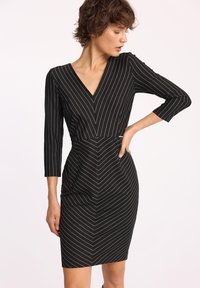 Czarna sukienka z rękawami o długości trzech czwartek, z dekoltem w kształcie litery V i dopasowanym krojem. Posiada białe pionowe paski w wzór chevron.