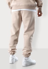 Beige Jogginghosen mit elastischen Bündchen, aus weichem Stoff, kombiniert mit weißen Sneakern. Minimalistisches Design mit einem dezenten Logodetail.