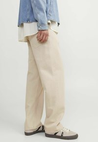 Jeans beige de pierna ancha con un acabado texturizado, combinados con una chaqueta de denim azul claro y una camisa a rayas debajo, luciendo con zapatillas deportivas.
