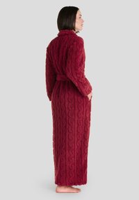 Bata de baño larga roja hecha de tela texturizada que presenta un patrón de punto trenzado, un collar y un cinturón a la altura de la cintura, con un diseño de longitud completa.