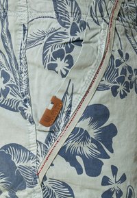 Shorts en tissu vert clair avec un motif floral bleu marine, dotés d'une fermeture éclair latérale avec des accents rouges et d'une étiquette de marque en cuir beige.