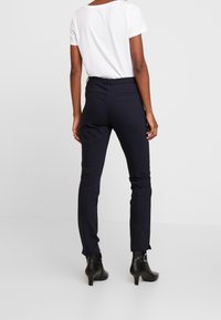 Pantalon slim-fit bleu marine avec une taille haute, texture lisse, poches arrière et design longueur cheville, associé à un haut à manches courtes blanc.