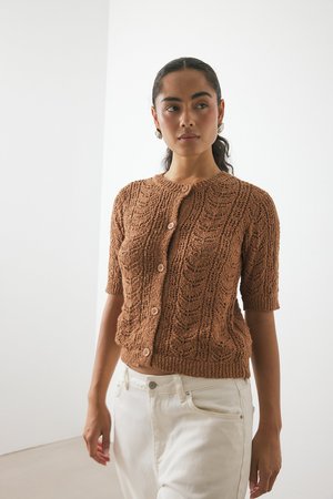 Femme portant un cardigan court en tricot marron à manches courtes avec des boutons, associé à un pantalon blanc taille haute, debout contre un mur blanc.