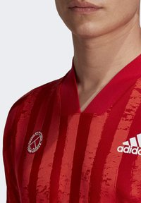 Jersey atlético rojo con rayas horizontales texturizadas. Presenta un cuello en V, un pequeño logo de Adidas y una insignia circular en el lado izquierdo.
