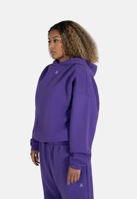 Lilla hettegenser med avslappet passform, laget av myk stoff, med en liten logo på brystet og matchende joggebukser.