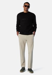 Sudadera negra con el logo "NORTH SAILS", pantalones beiges y zapatillas verdes. El modelo tiene el pelo corto y lleva gafas de sol oscuras.