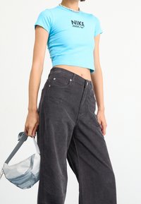 Personne portant un t-shirt court Nike bleu vif, un pantalon large noir, un collier en argent et tenant un sac banane gris clair North Face.