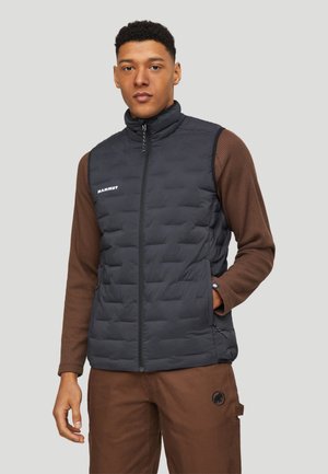 Ung mand iført en sort quiltet vest over en brun sweater og brune bukser med Mammut-logoer, stående med den ene hånd i lommen.