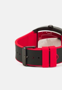 Bracelet de montre en silicone noir et rouge avec une finition mate, comportant une boucle métallique et des trous pour la boucle. L'arrière montre un boîtier transparent.