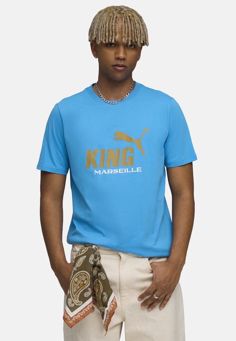 Šviesiai mėlynas medvilninis marškinėlis su auksiniais užrašais "KING" ir "MARSEILLE", dekoruotas Puma logotipu. Ruda paisley tipo skarelė užrišta ant liemens.