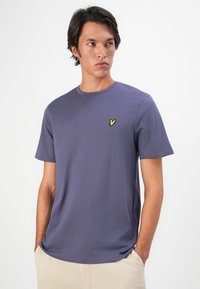Joven vestido con una camiseta morada lisa con un pequeño logo de un pájaro amarillo en el lado izquierdo del pecho, de pie con las manos en los bolsillos de un pantalón beige.
