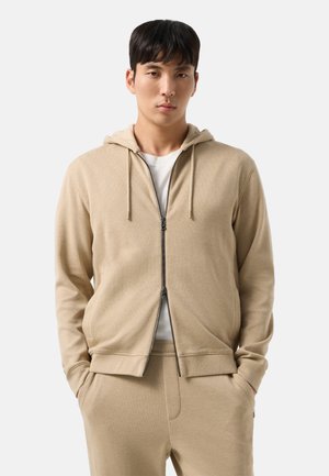 Jeune homme aux cheveux noirs courts portant un sweat à capuche beige zippé sur une chemise blanche et un pantalon beige assorti, mains dans les poches, expression neutre.